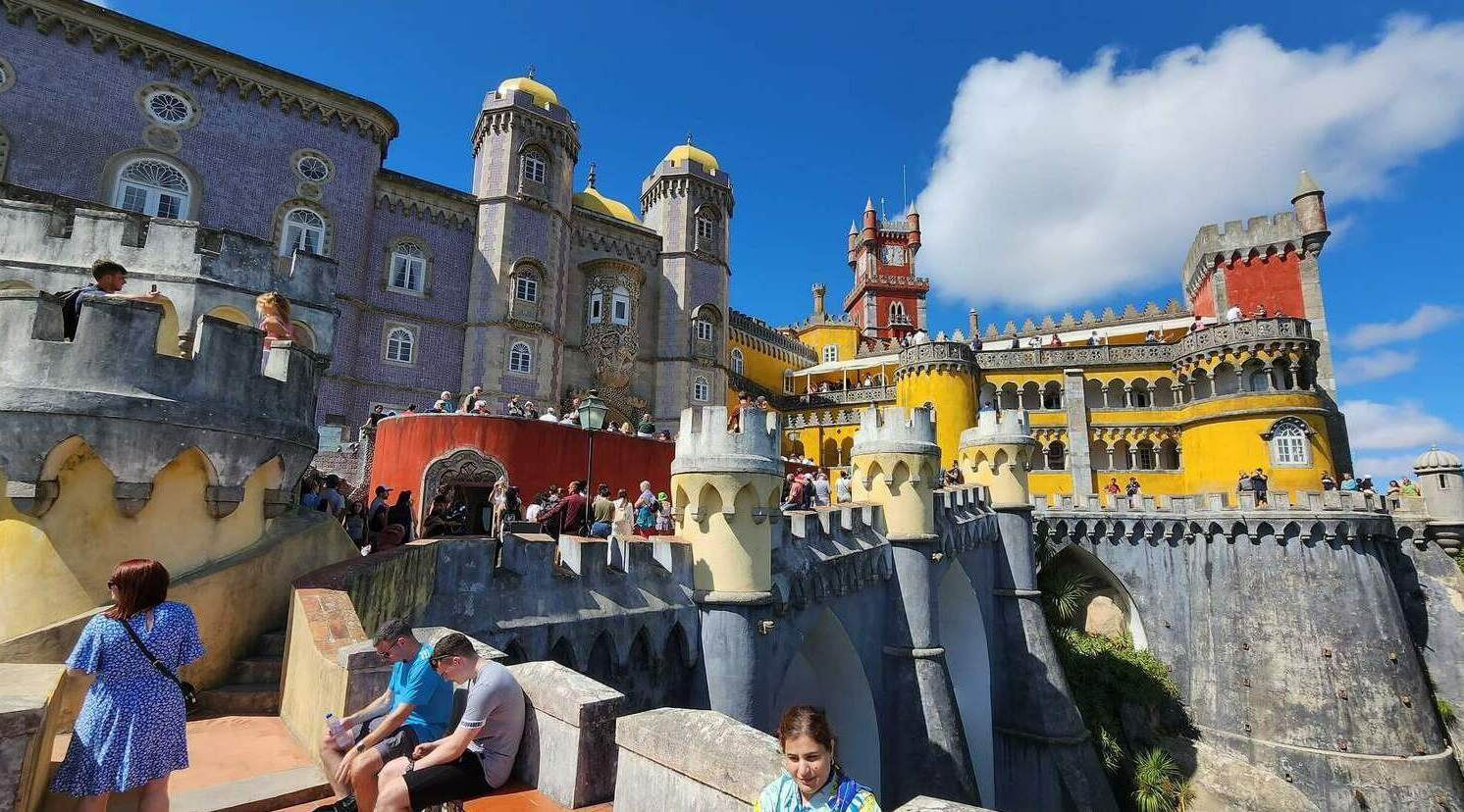 Яркие желтые и красные башни дворца Пена в Синтре, вид с террасы с туристами под ярко-синим небом.