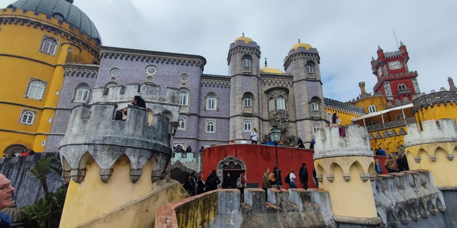 Разноцветные фасады дворца Пена в Синтре с желтыми башнями и фиолетовыми стенами под облачным небом.