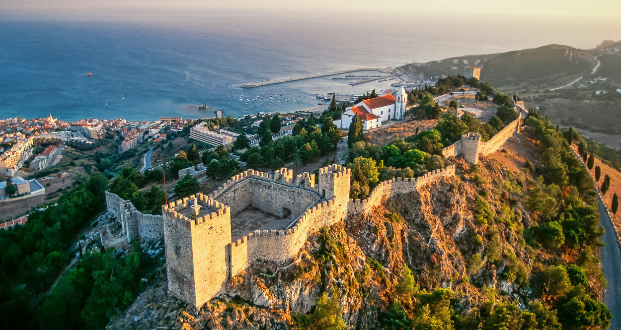 Вид с высоты на мавританский замок Сезимбра на закате: крепостные стены, город у подножия и панорама Атлантического океана.